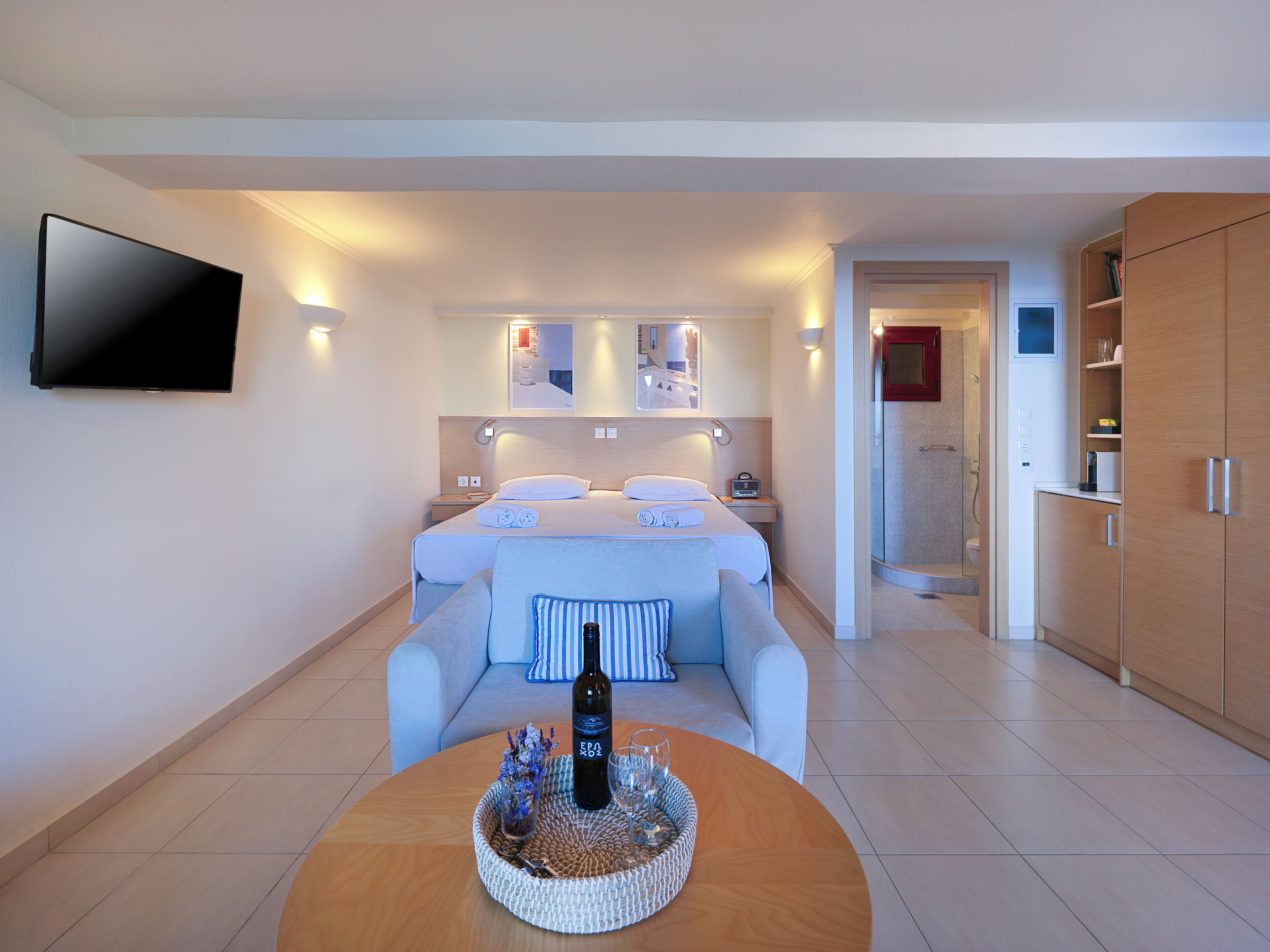 Una habitació d'hotel moderna amb un llit gran, un televisor fixat a la paret, una taula petita amb una ampolla i vasos, i un plat de dutxa obert.