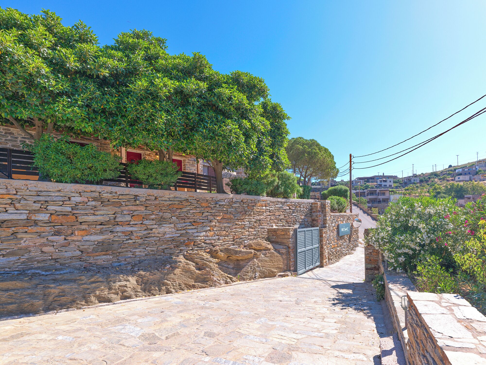 A stone pathway leading to a stone wall with a gate, surrounded by greenery and buildings in the background.