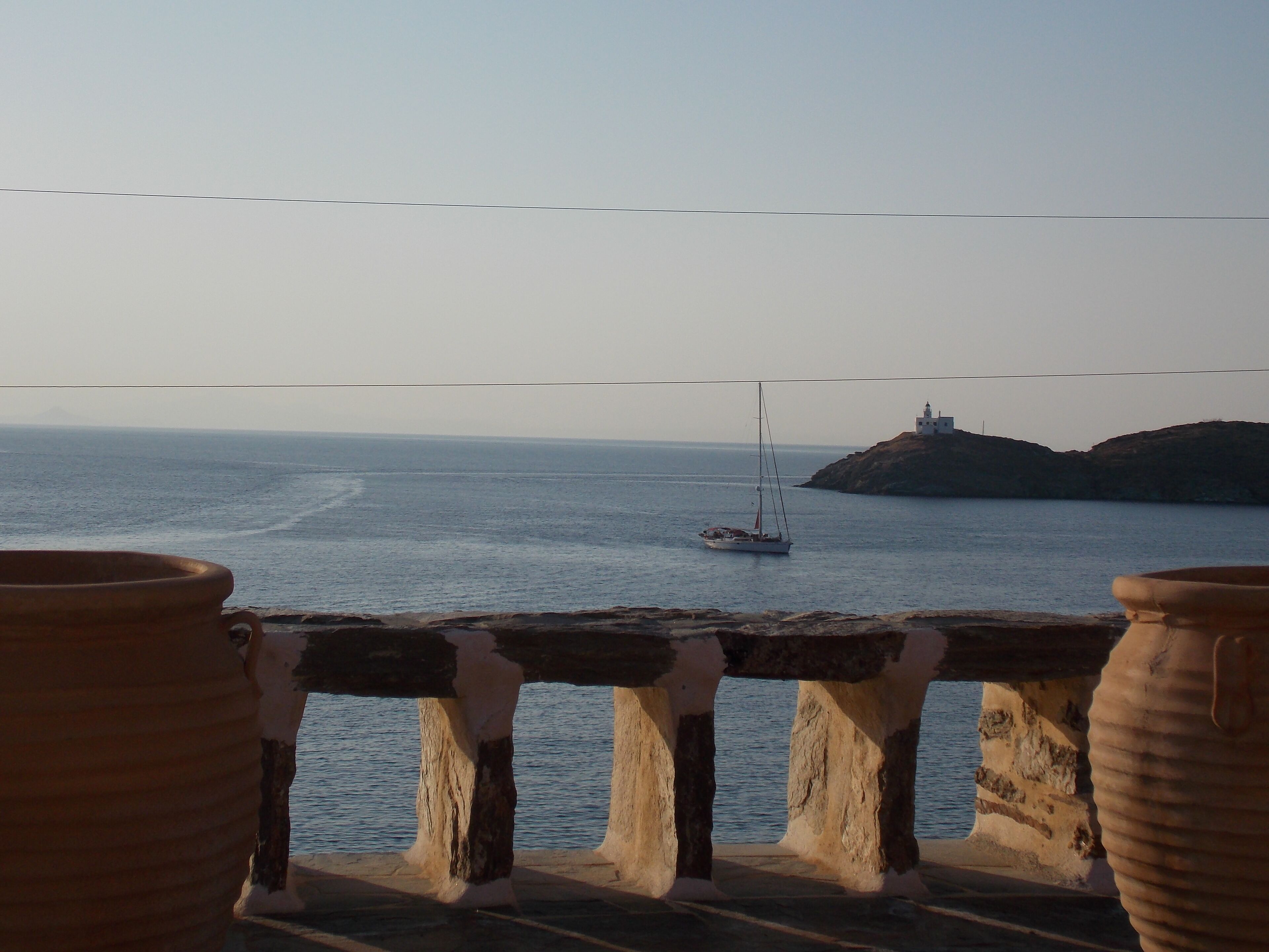 A coastal view with a sailboat on the water, a lighthouse on a hill, and large terracotta pots in the foreground.