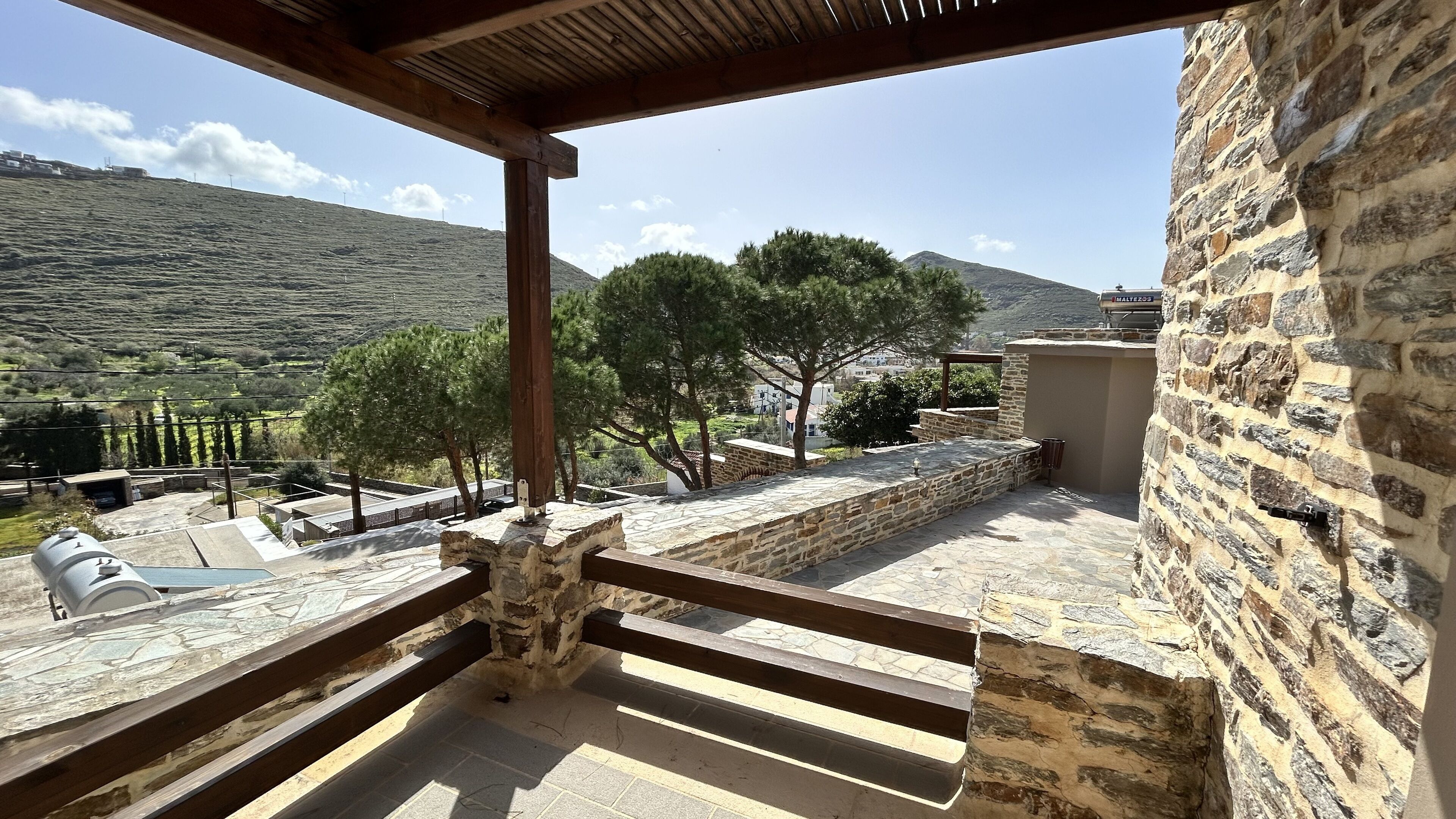 A balcony with stone walls, a wooden railing, and a view of a hilly landscape with trees and a distant mountain.