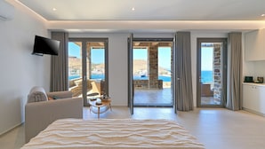 A hotel room with a large bed, a sofa, a TV, and a view of the sea through open doors.