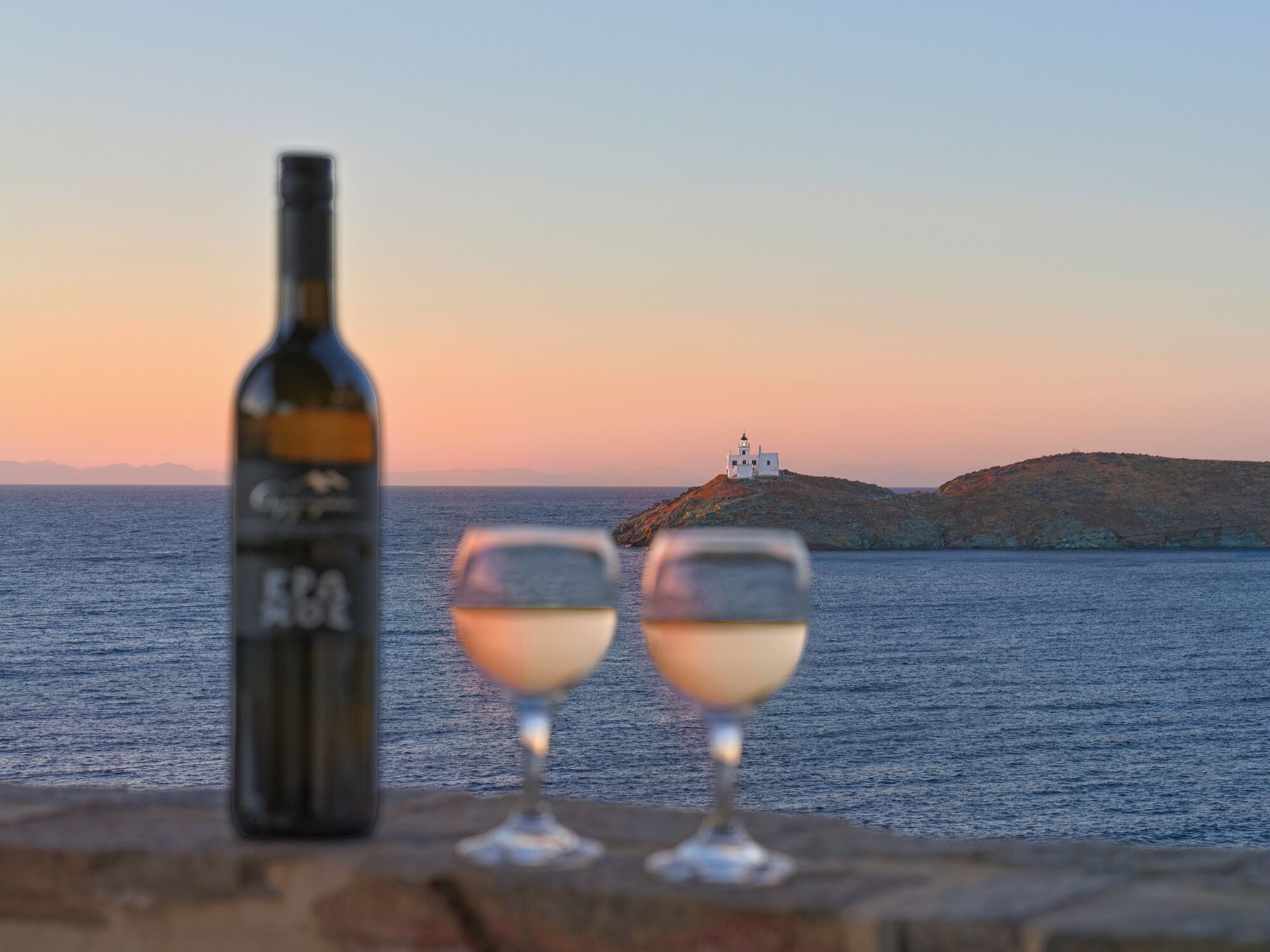 A bottle and two glasses on a ledge with a view of the sea and a lighthouse in the distance.