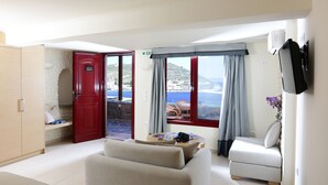 A modern hotel room with a bed, a sofa, a small table, and a TV. The room has a view of the sea and a red door.