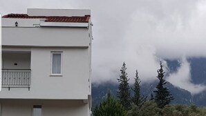 A white building with a balcony and a window, surrounded by trees and mountains in the background.