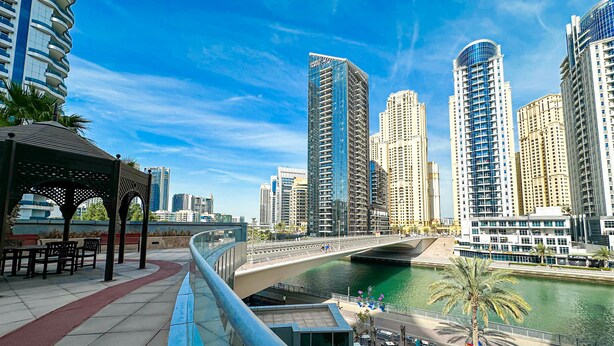 A riverside walkway with outdoor seating, a bridge, and high-rise buildings in the background.
