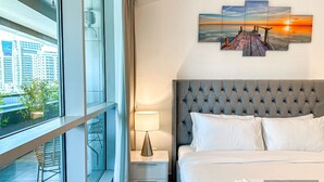A hotel room with a large window, a bed with white linens, a grey tufted headboard, a bedside table with a lamp, and a sunset view on the wall.