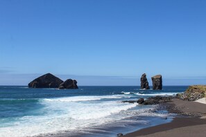 A beach with dark sand, waves, and two prominent rock formations in the ocean. - Casa Pé na Areia (Ponta Delgada)