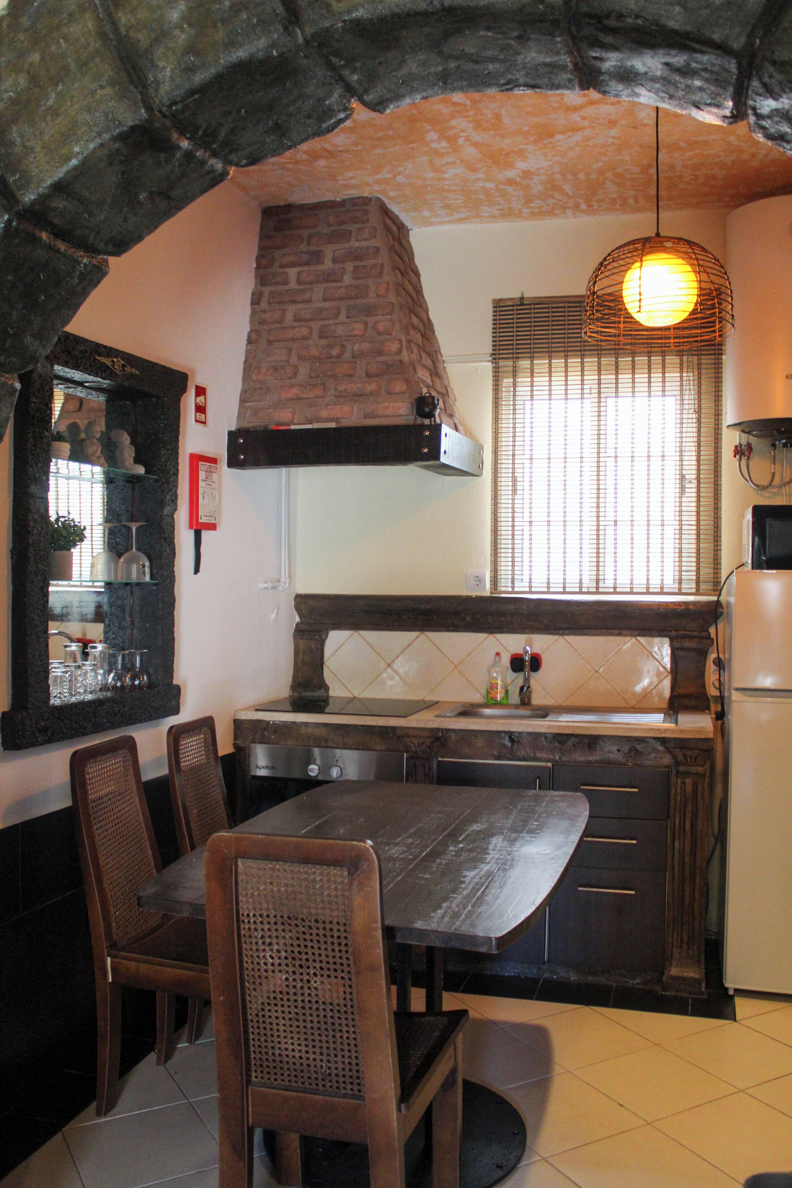 Una cocina con pared de ladrillo, lámpara colgante, ventana con persianas y refrigerador.