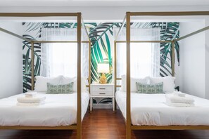 A room with two beds, each with white bedding and pillows, a nightstand with a lamp, and a tropical leaf-patterned wall.