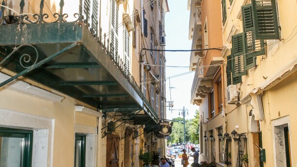 A narrow street with buildings on either side, a scooter parked on the right, and a person walking in the distance. - Conte Studio (Corfu)