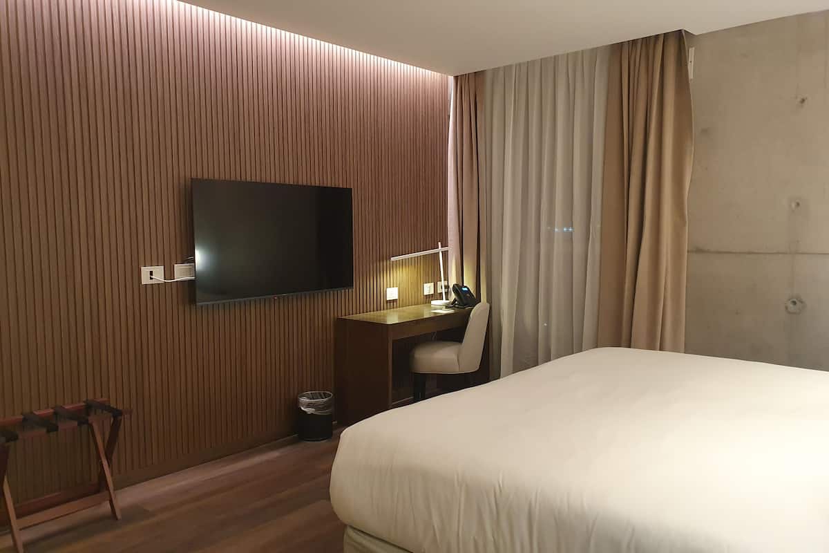 Une chambre d’hôtel avec un grand lit, un bureau avec une chaise, un téléviseur fixé au mur et une fenêtre avec des rideaux.