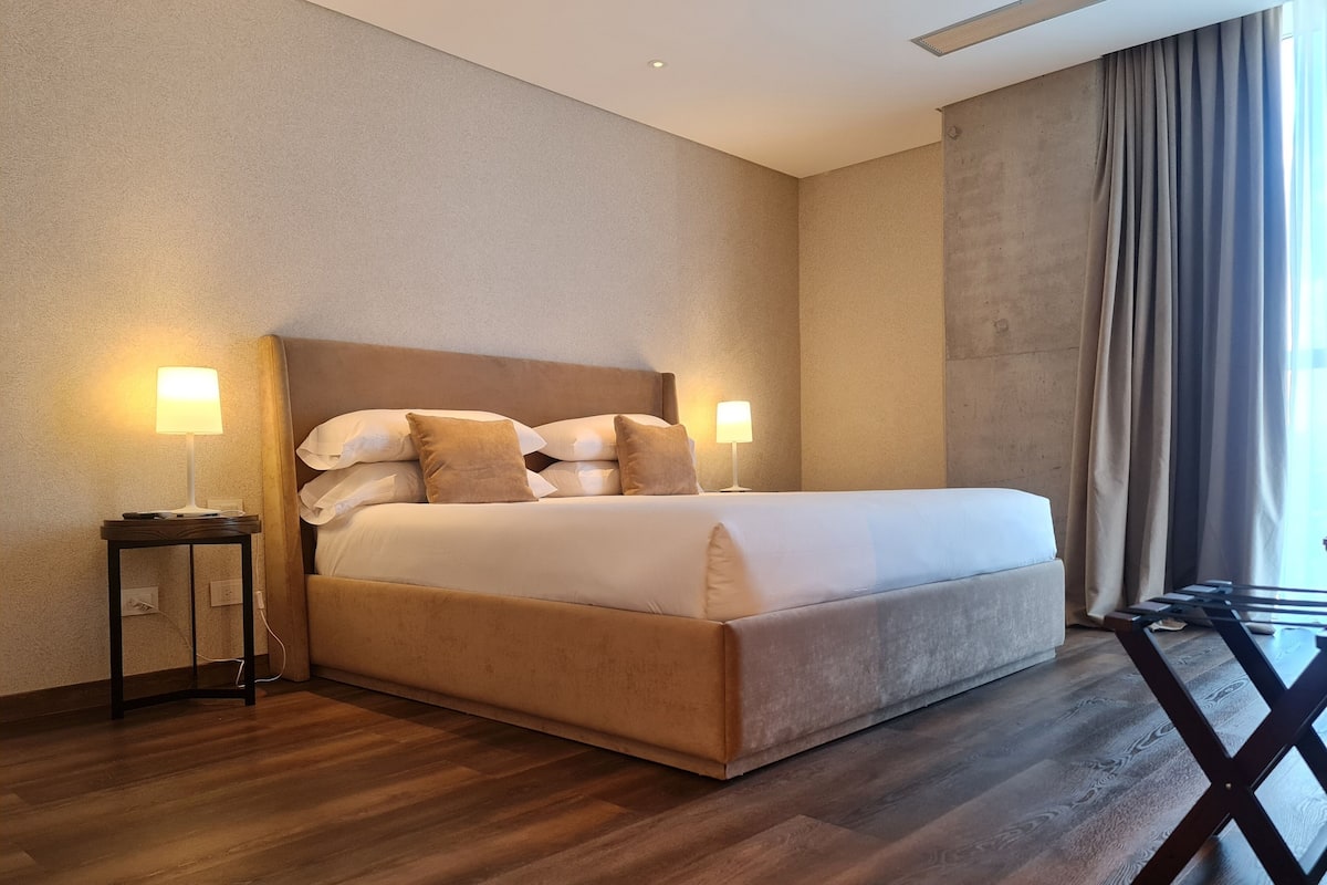 Une chambre d’hôtel moderne avec un grand lit, deux lampes de chevet, un plancher en bois et une vue sur l’extérieur par une fenêtre munie de rideaux.