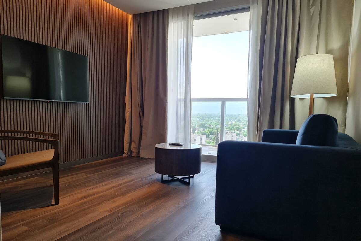 Une chambre d’hôtel moderne avec un plancher en bois, un téléviseur à écran plat fixé au mur, un canapé bleu foncé, une chaise en bois, une petite table ronde et une grande fenêtre avec des rideaux.