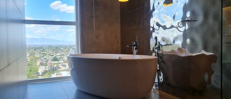 A modern bathroom with a freestanding bathtub, a large window offering a city view, and a unique textured wall.