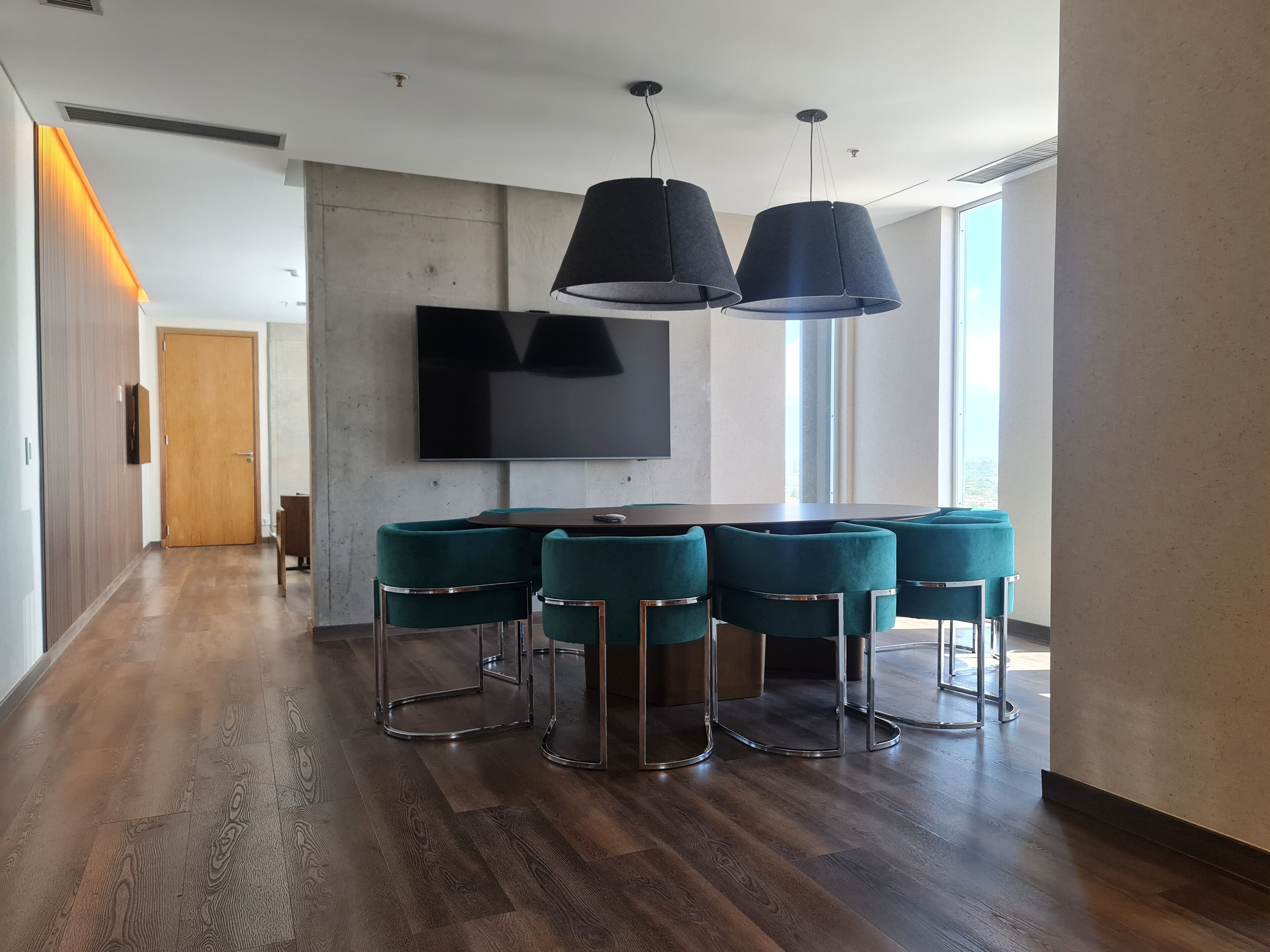 A modern dining area with a round table and six teal chairs, a flat-screen TV on the wall, and a large window providing natural light.