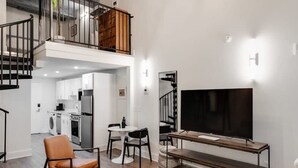 En moderne stue med sofa, sofabord, fjernsyn og et spiseområde med stole og bord. Et køkken ses i baggrunden.