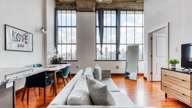 A modern living room with a long grey sofa, a dining table with chairs, a TV on a wooden cabinet, and large windows with a city view. - Sosuite 2BR Penthouse w W D Gym On-site Bar (Philadelphia)