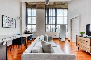 A modern living room with a long grey sofa, a dining table with chairs, a TV on a wooden cabinet, and large windows with a city view. - Sosuite 2BR Penthouse w W D Gym On-site Bar (Philadelphia)