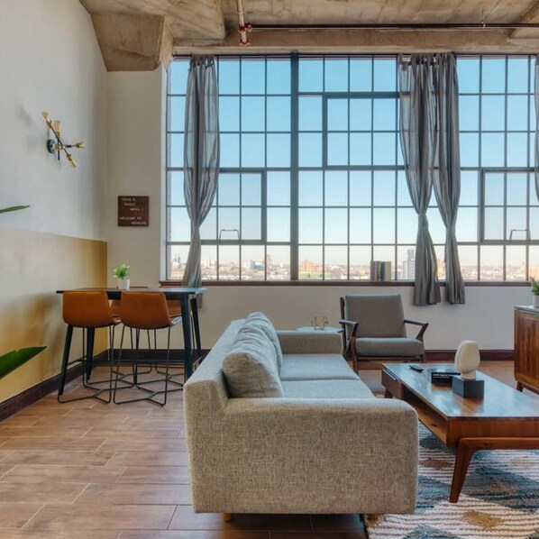A modern living room with a sofa, chairs, a coffee table, and a dining area with a table and chairs. Large windows with curtains provide a view of the cityscape.
