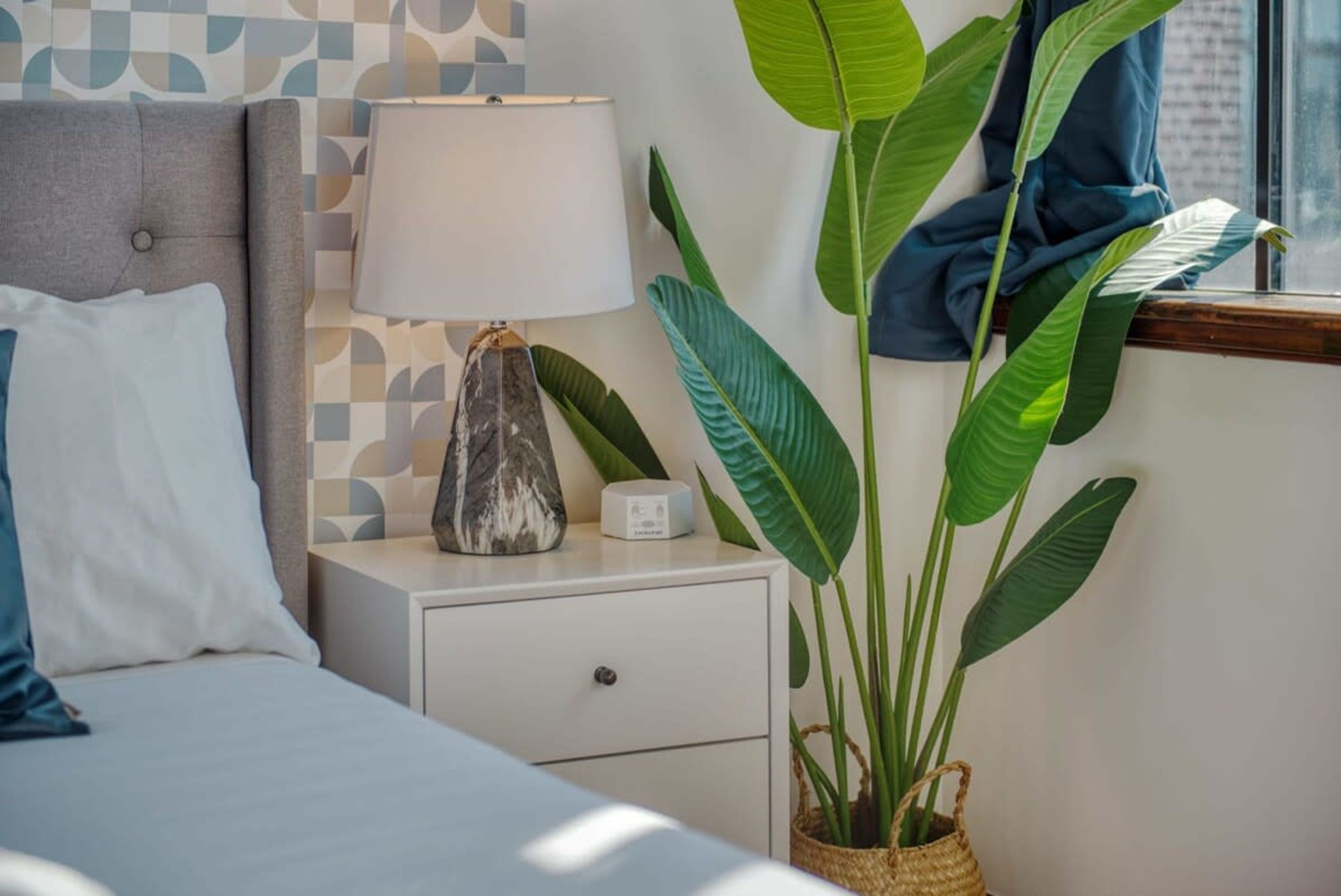 A neatly made bed with pillows, a bedside lamp, a small bedside table with a decorative vase, and a large potted plant.