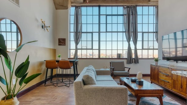 A modern living room with a sofa, dining table, chairs, a TV, and large windows with curtains. - Sosuite 2BR Penthouse w Balcony W D Gym (Philadelphia)