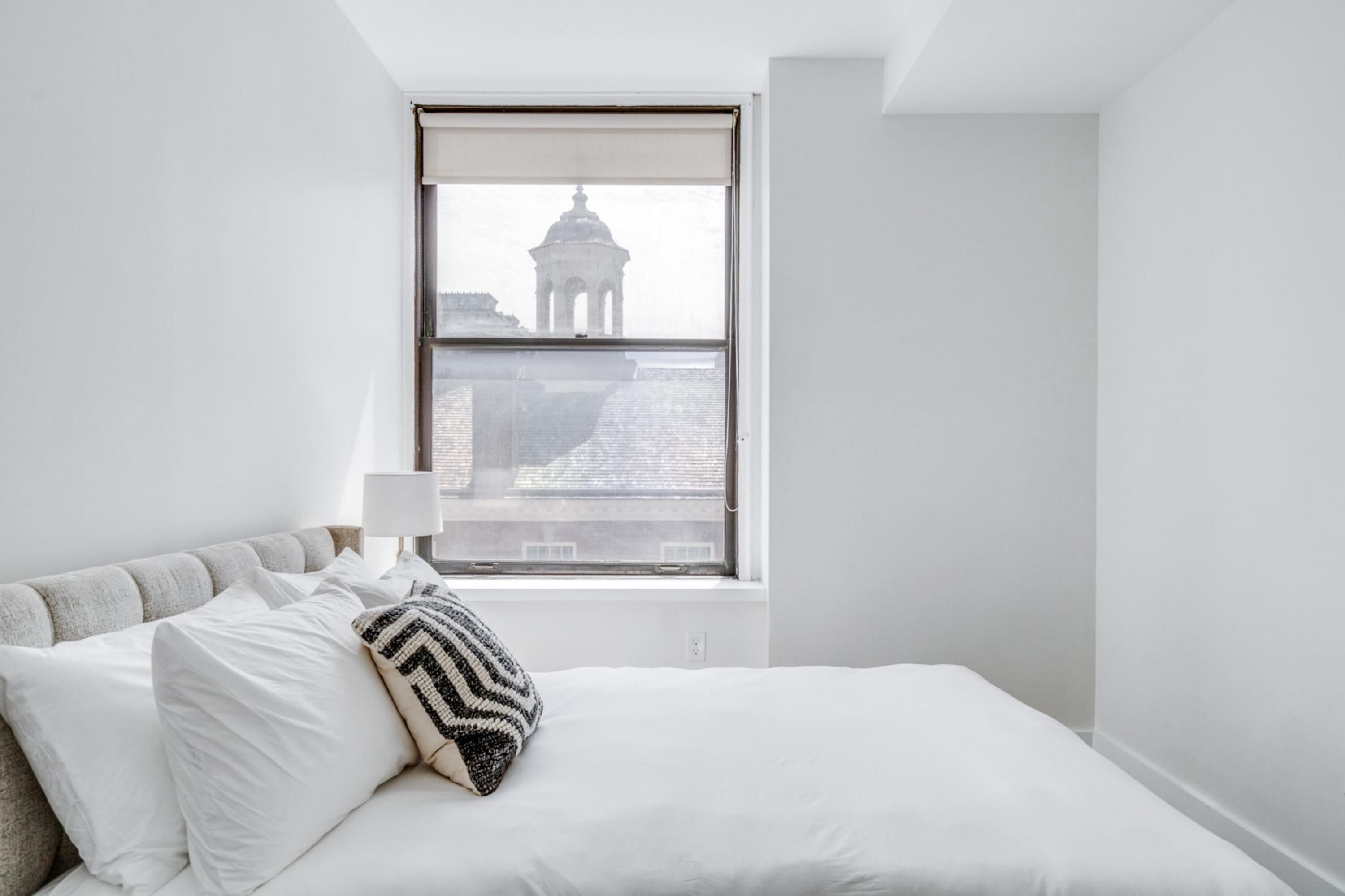 A minimalist bedroom with a large window, a bed with white bedding, and a patterned throw pillow.