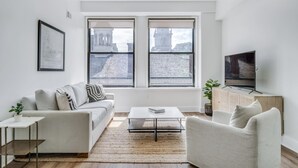 A modern living room with a sofa, coffee table, and TV, large windows with a city view, and a framed picture on the wall. - Sosuite 2BR Apt w Park View, W/d, Gym, Lounge (Philadelphia)