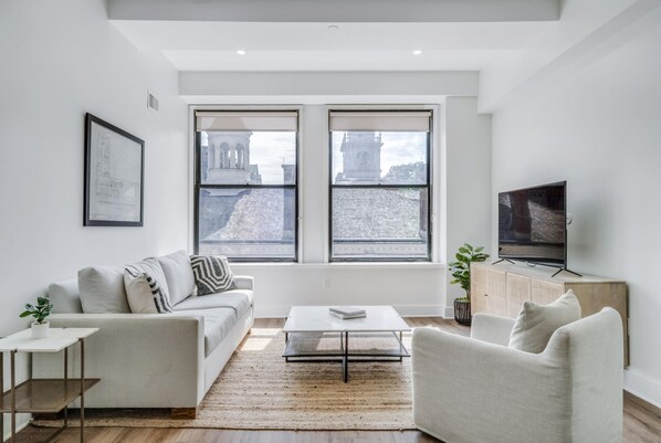 A modern living room with a sofa, coffee table, and TV, large windows with a city view, and a framed picture on the wall.