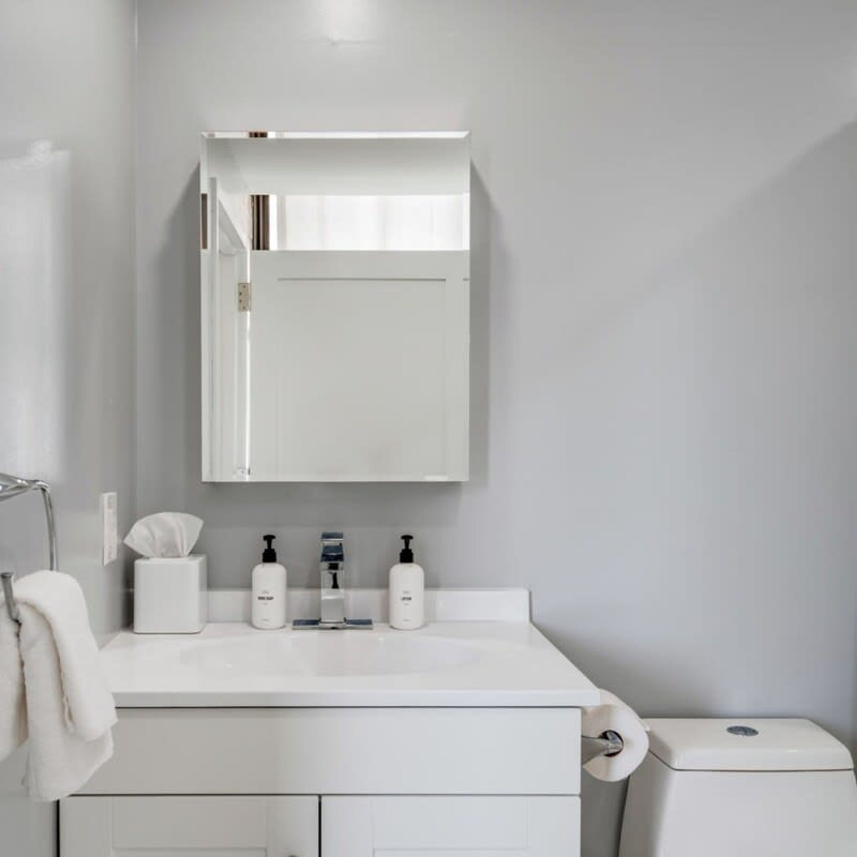 A modern bathroom with a white sink, mirror, and toilet. There are three bottles of hand soap and a tissue box on the sink.