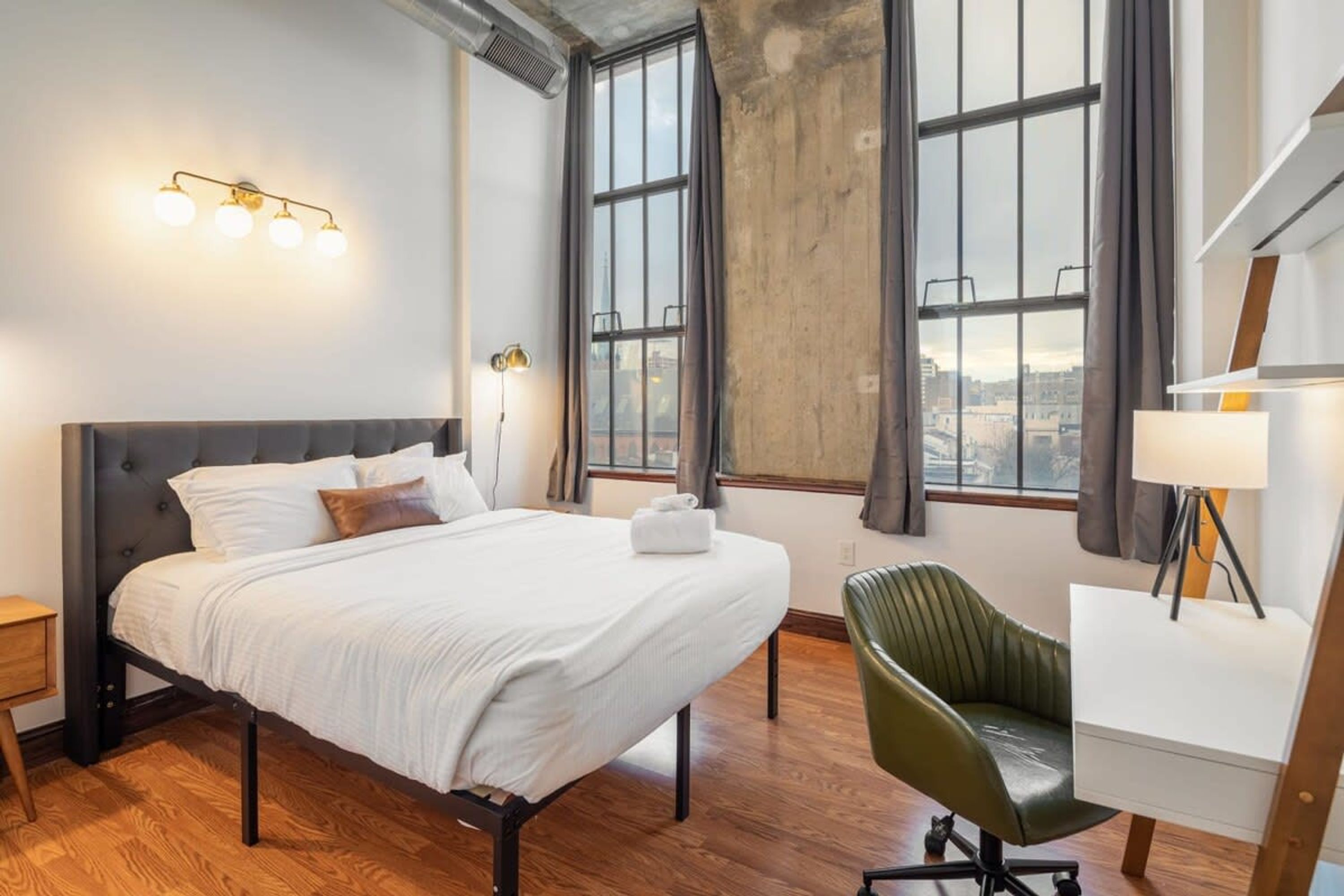 Camera d'albergo con un letto, una scrivania e una sedia. Sono presenti ampie finestre con tende, una lampada e un punto luce a parete.