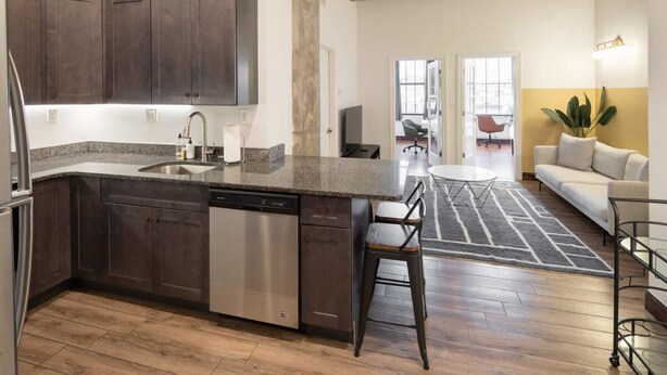 A modern kitchen with dark cabinets, a dishwasher, and a granite countertop. There is a dining area with a round table and chairs, and a sofa in the background. - Sosuite 2BR Apt w W/d, Gym, On-site Restaurant (Philadelphia)