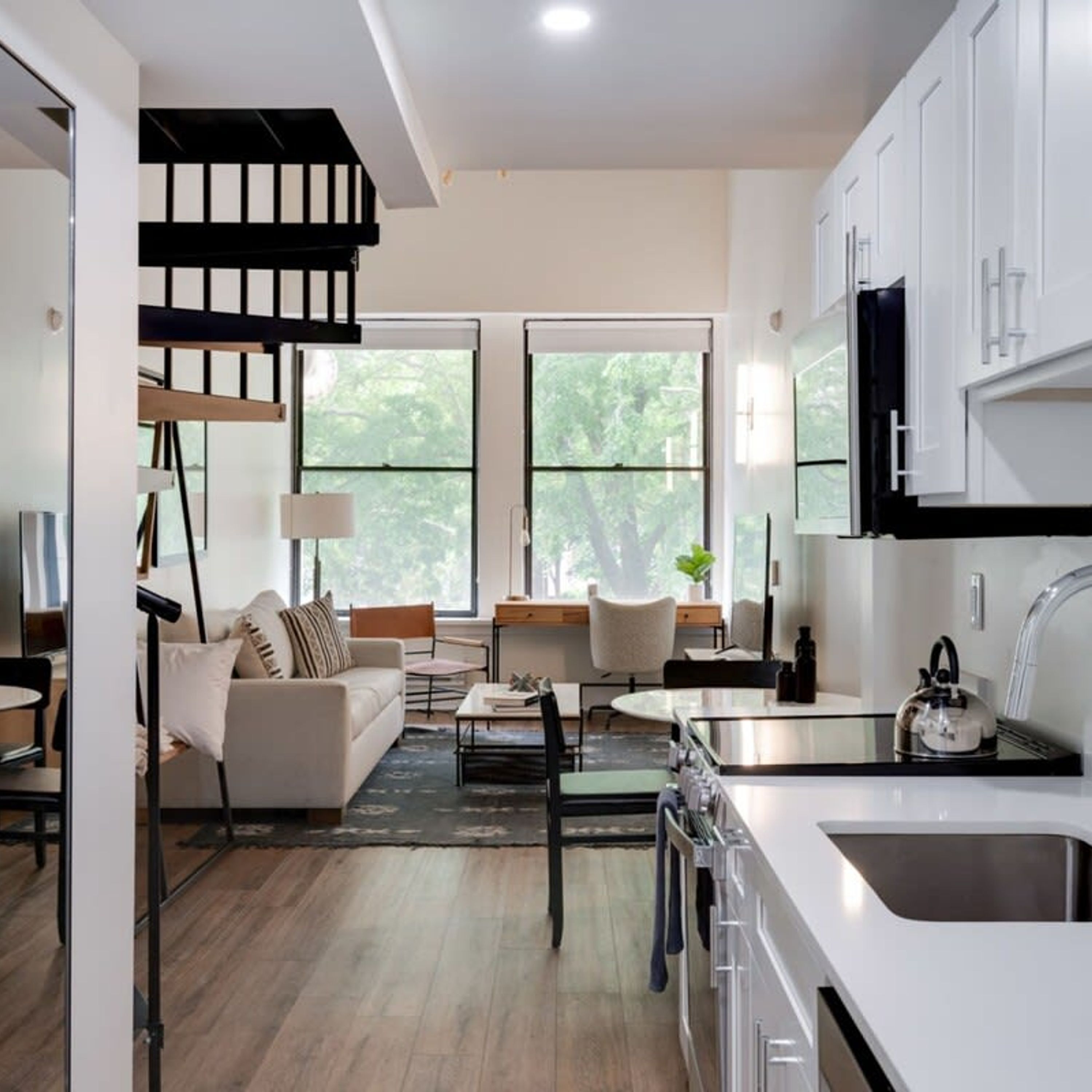 A modern kitchen with white cabinetry, a dining area with a sofa and coffee table, and a staircase with black railings.
