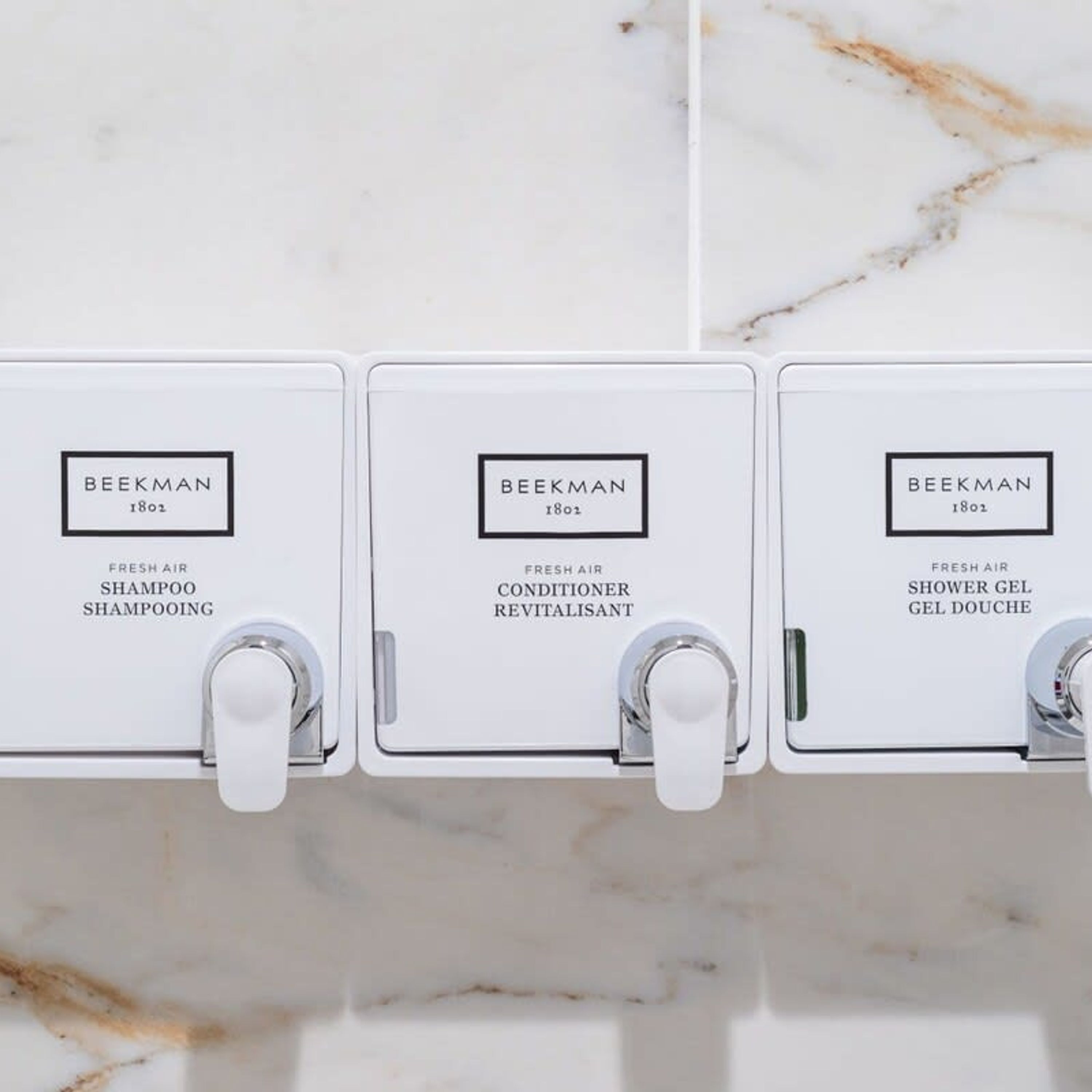 Three white dispensers labeled 'Beekman 1802' for fresh air shampooing, conditioner revitalisant, and shower gel gel douche.