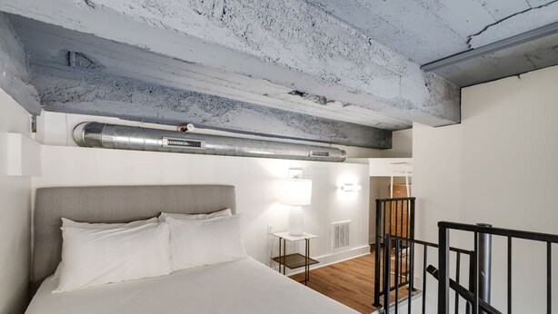A bedroom with a large bed, a small table with a lamp, a radiator, and a staircase with a railing. - Sosuite 1BR Loft w Shared W/d, Gym, Lounge (Philadelphia)