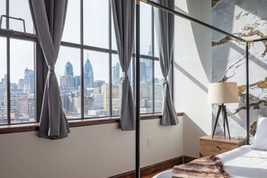 A hotel room with a large window offering a city skyline view, a bed with a white comforter, a bedside table with a lamp, and a marble-patterned wall. - Sosuite 2BR Penthouse w W D Gym On-site Bar (Philadelphia)