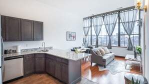 A modern kitchen with dark cabinets, a marble countertop, and a dishwasher. There is a dining area with a gray sofa and a city view through large windows. - Sosuite 2BR Penthouse w W D Gym On-site Bar (Philadelphia)