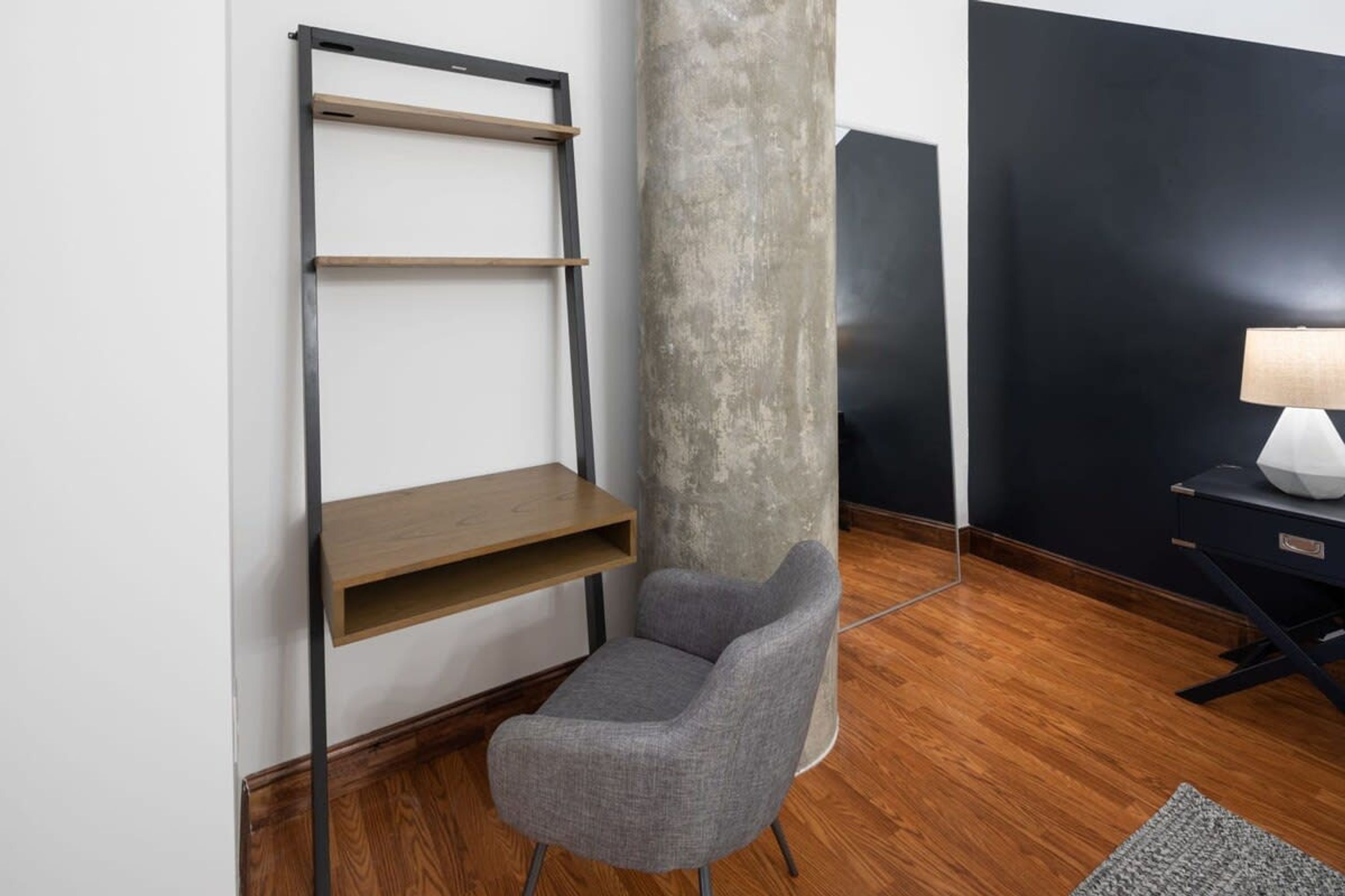 Una habitación moderna con un sillón gris, una estantería de madera, un pilar de hormigón y un escritorio con una lámpara.