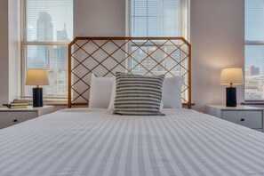 Quarto de hotel com uma cama, duas luminárias de cabeceira, uma janela com persianas e vista para a cidade.