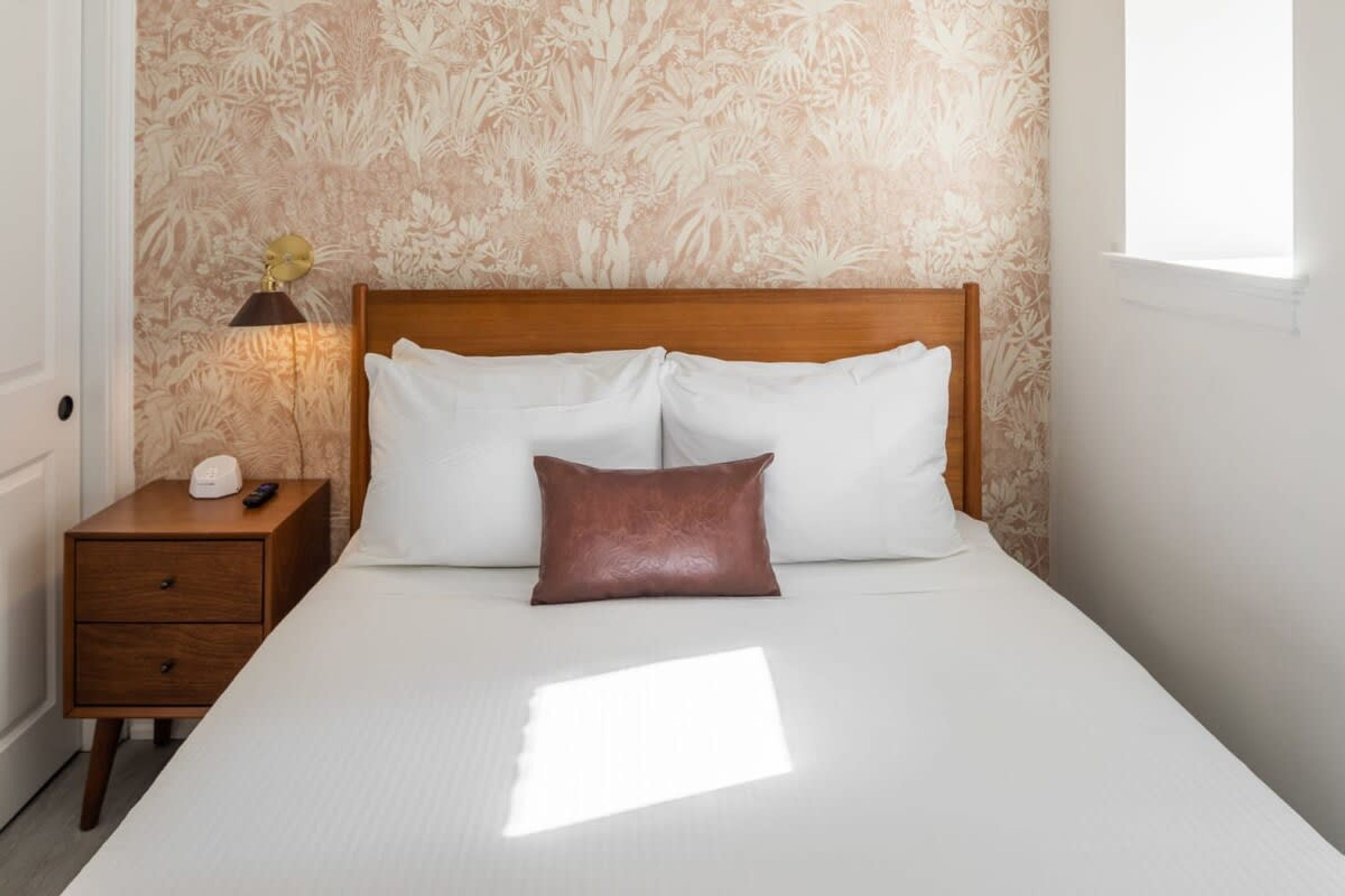 A neatly made bed with white linens and a single brown pillow, a wooden nightstand with a lamp, and a floral wallpaper pattern.