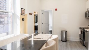 A modern kitchen with a dining area, a trash can, and a view of the city through a large window. - Sosuite 1BR Apt w W/d, Large Dining Seating (Philadelphia)