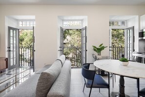 Ein modernes Wohnzimmer mit einer grauen Couch, einem runden Tisch mit Stühlen und einem Balkon mit Blick auf Bäume.