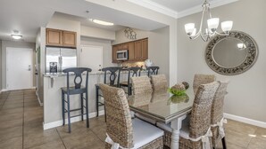 A dining area with a glass table, wicker chairs, a chandelier, and a decorative mirror. - Gulf Front with Beach Views & Large Balcony (Miramar Beach)