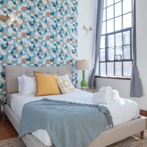 A neatly made bed with a blue throw blanket, a yellow pillow, and white bedding. There is a green lamp on a small table beside the bed, and a window with curtains to the right. - Sosuite 2BR Penthouse w Balcony W D Gym (Philadelphia)