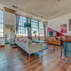 Ein geräumiges Wohnzimmer mit einer Couch, einem Essbereich mit Tisch und Stühlen, großen Fenstern mit Vorhängen und Blick auf die Stadt.