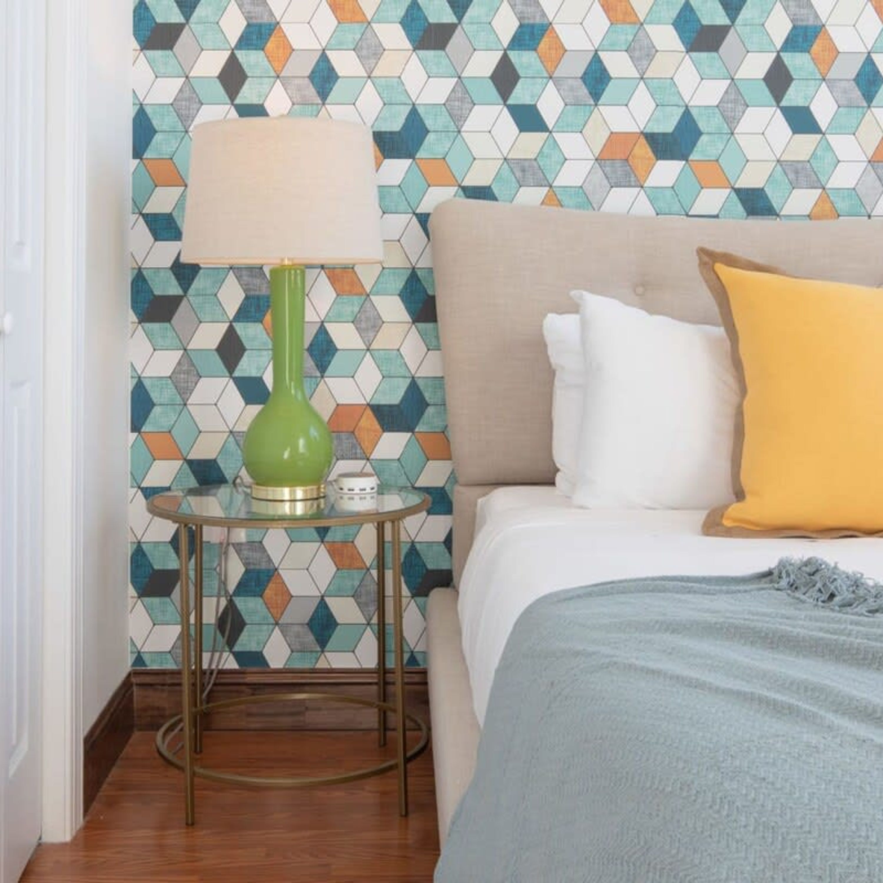 A hotel room with a patterned wall, a bed with white and yellow pillows, a green lamp on a small table, and a blue throw blanket.