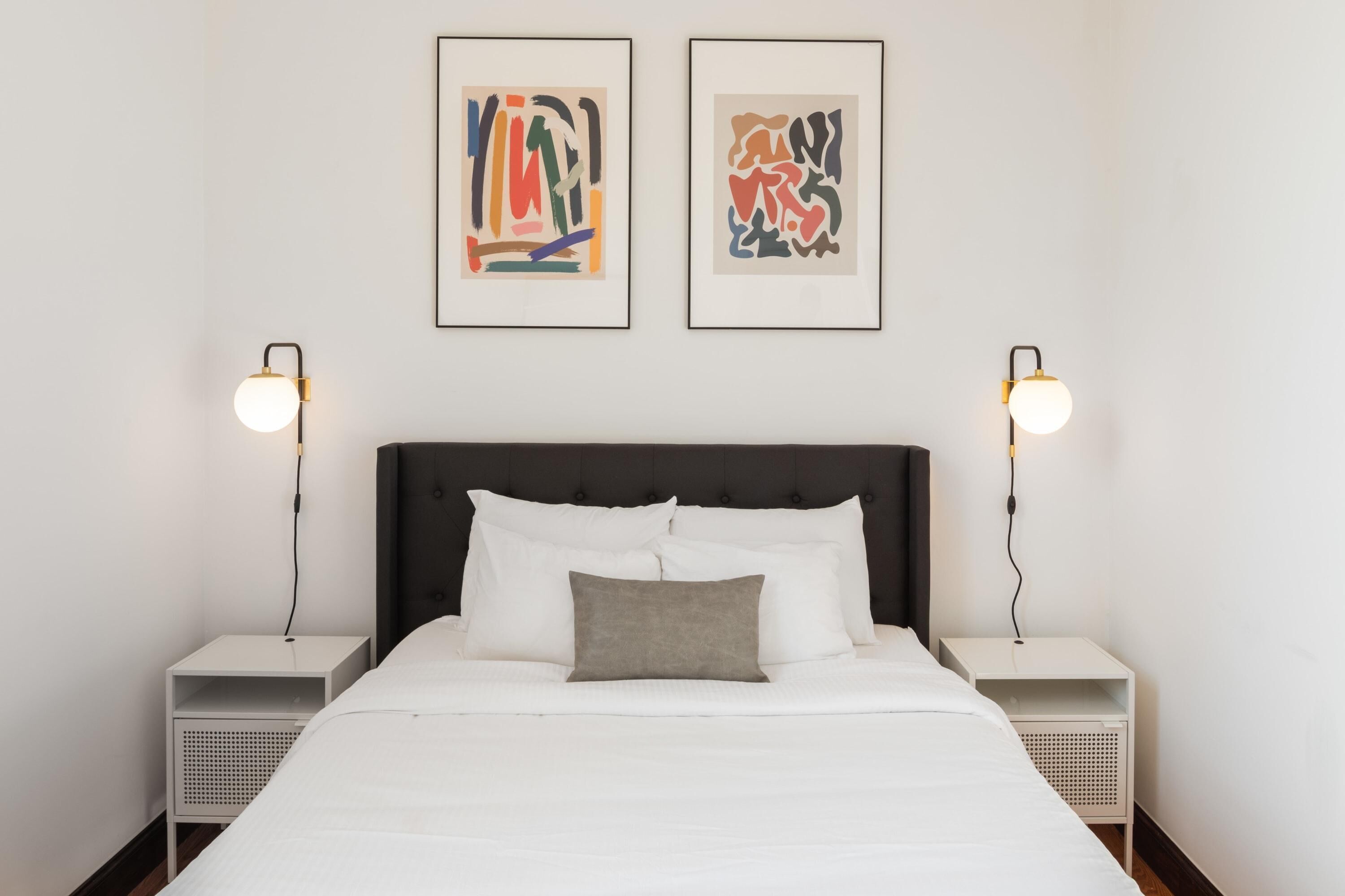 A neatly made bed with white linens and a gray pillow, flanked by two bedside tables with lamps, and two framed abstract artworks on the wall.