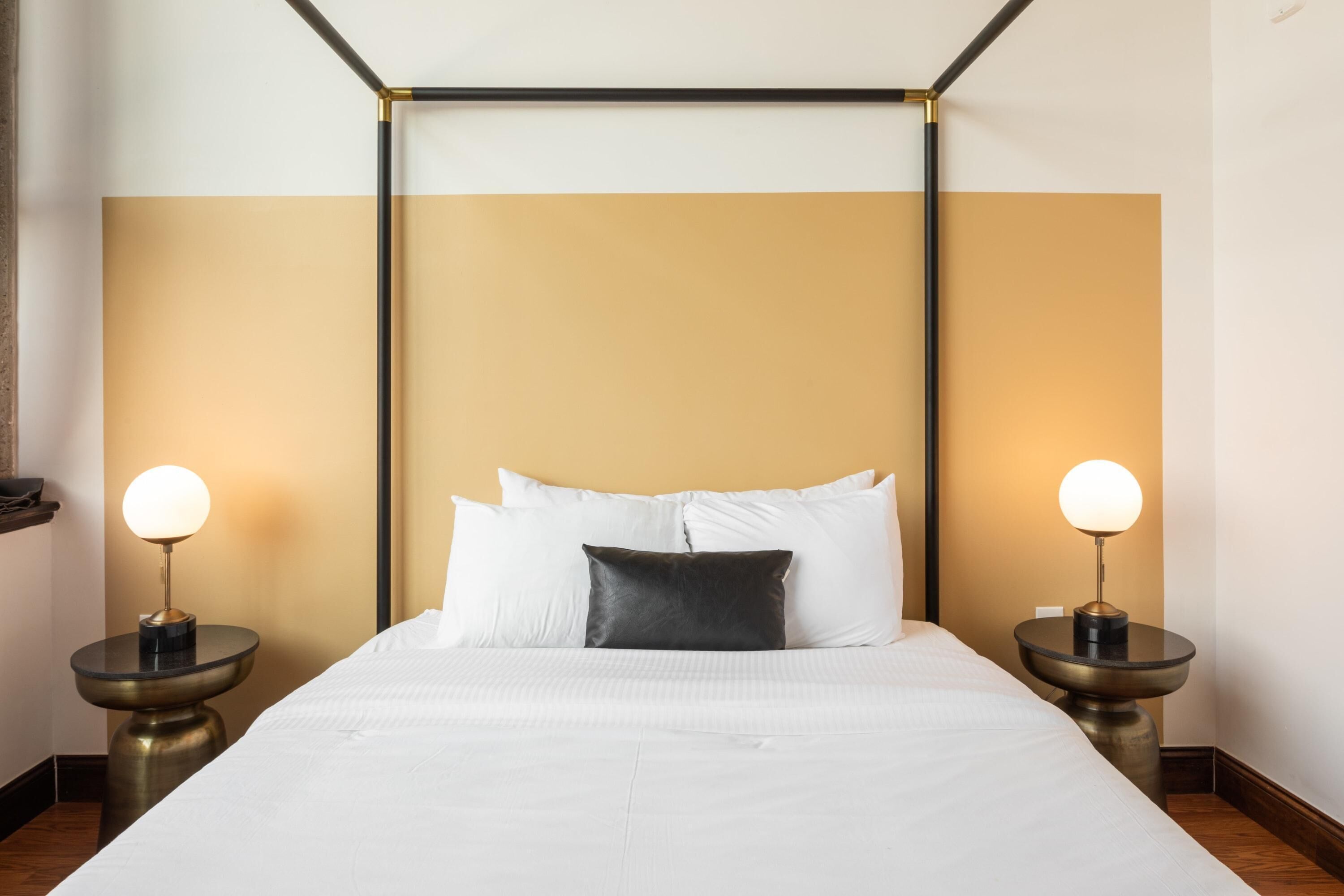 Un lit bien fait, avec du linge de lit blanc et un oreiller foncé, flanqué de deux tables de chevet surmontées de lampes, dans une pièce au design simple et épuré.
