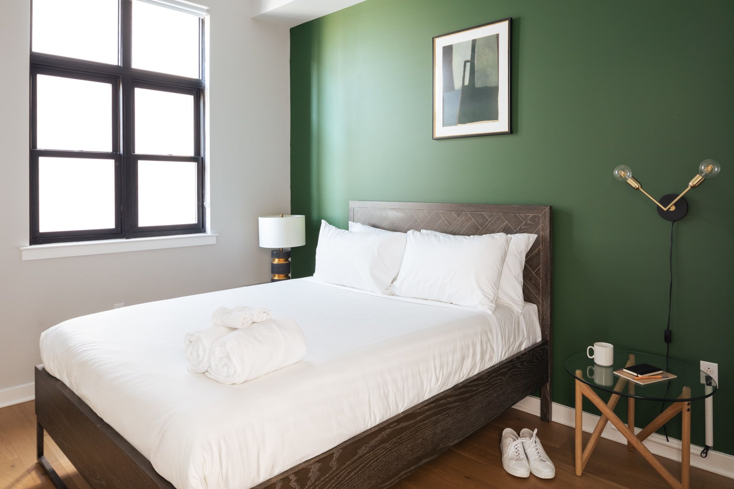 A neatly made bed with white linens and a rolled towel, a bedside table with a lamp, a framed picture on the wall, and a small table with a book and a cup.