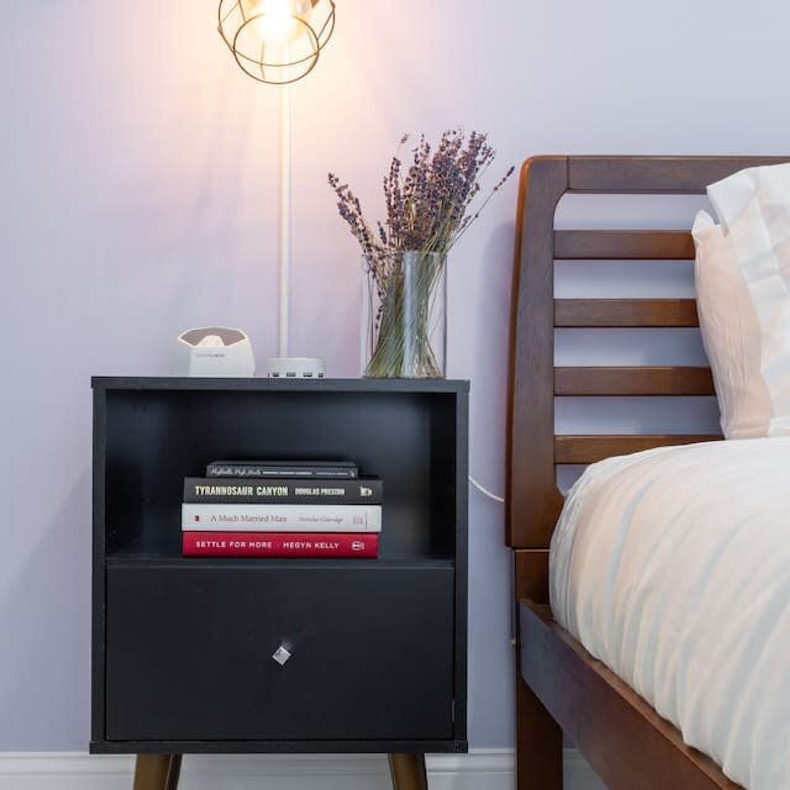 Una cama bien hecha con cabecera de madera, una mesita de noche con lámpara y un jarrón con flores secas.