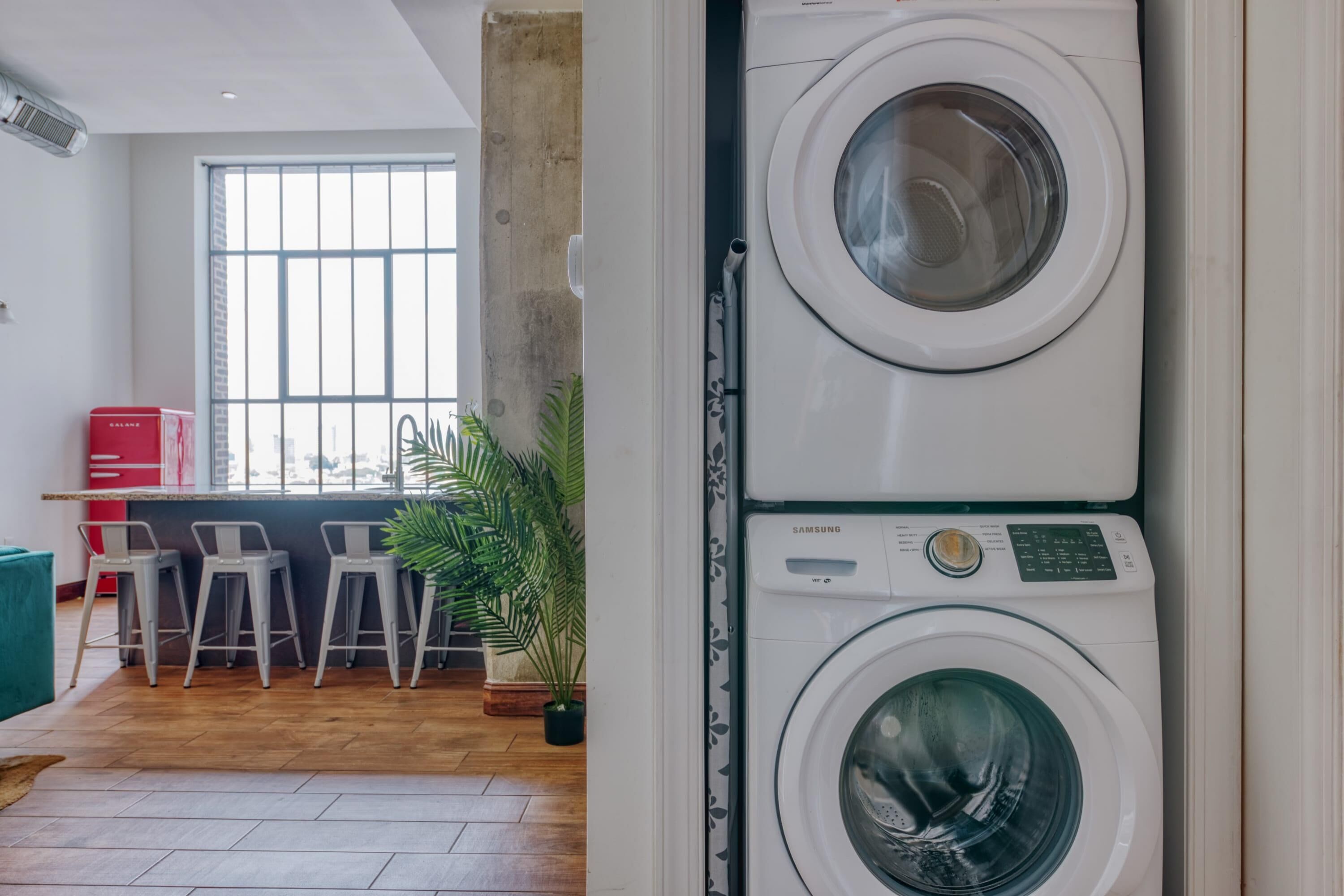 A laundry area with stacked washing machines, a bar counter, and seating area with chairs and a potted plant.